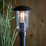 Chartley Outdoor Post Light in textured black aluminium with clear polycarbonate shade, lit E27 bulb visible, set against a wooden fence and foliage.