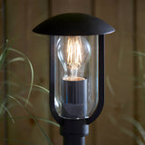 Black textured aluminium Chartley Outdoor Post Light with domed canopy and clear polycarbonate shade housing a lit E27 bulb, set against a garden backdrop.