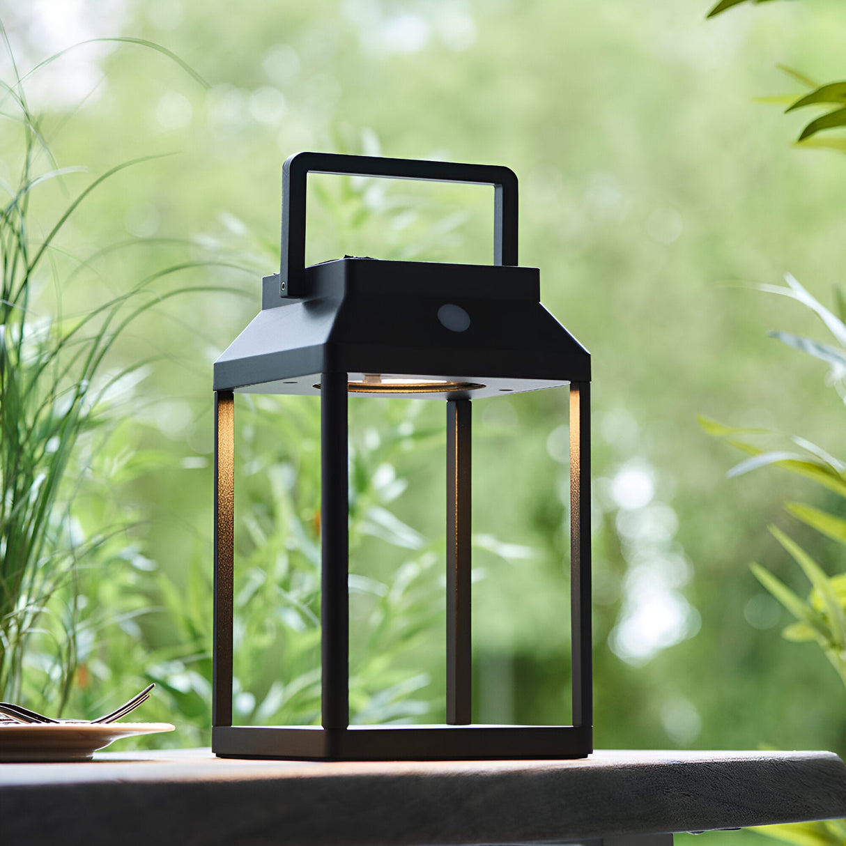 Caldshaw LED Solar Table Lamp in black, lantern-style aluminium frame with warm white glow, shown outdoors on a table amid greenery.