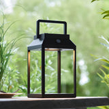 Caldshaw LED Solar Table Lamp in black, lantern-style aluminium frame with warm white glow, shown outdoors on a table amid greenery.