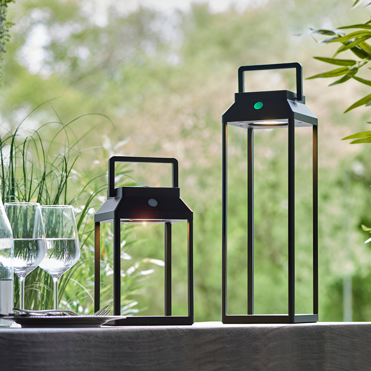 Caldshaw LED Solar Table Lamps in black, lantern-style frames with carry handles shown outdoors on a table beside wine glasses and greenery.