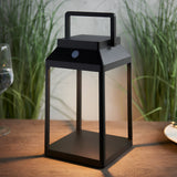 Caldshaw LED Solar Table Lamp in black, lantern-style aluminium frame with carry handle, shown lit on an outdoor wooden table beside greenery.