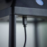 Close-up of the Caldshaw LED Solar Table Lamp in black showing the USB charging port and cable beneath the textured metal base.