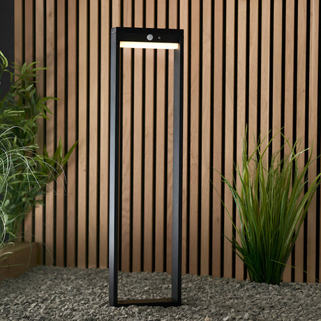 Endon Blythe LED solar bollard light in black aluminium with rectangular frame and PIR sensor, set on gravel against a wooden slatted wall with plants.