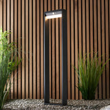 Endon Blythe LED solar bollard light in textured black aluminium with white diffuser and PIR sensor, set on gravel against a wooden slat wall with grasses.