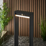 Endon Blythe LED solar bollard light in black aluminium with warm white diffuser and PIR sensor, shown on gravel against a slatted wood wall.