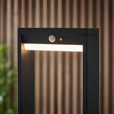 Endon Blythe LED solar bollard light in matte black aluminium with white diffuser and PIR sensor, illuminated outdoors against a ribbed brown backdrop.