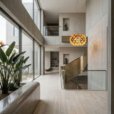 A modern, spacious interior with floor-to-ceiling windows, a brass staircase, light stone floors, indoor plants, minimalist decor, and contemporary lighting featuring the Briller LED Large Suspension Light in gold above the stairwell.