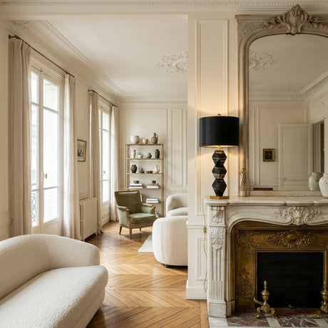 Elegant living room with classic French decor, gold-accented fireplace, ornate mirror, modern white sofas, herringbone floors, tall curtained windows, and a cozy reading nook illuminated by the Zerava 1 Light Faceted Marble Table Lamp - Black & Brass.
