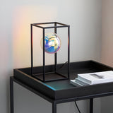 Brockwell Table Lamp in matt black with an open geometric frame and iridescent orb glass shade, shown lit on a black tray table beside books.