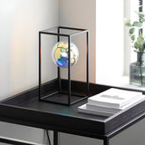 Brockwell Table Lamp in matt black, open rectangular frame with iridescent orb glass shade, shown on a bedside table beside books and a vase.