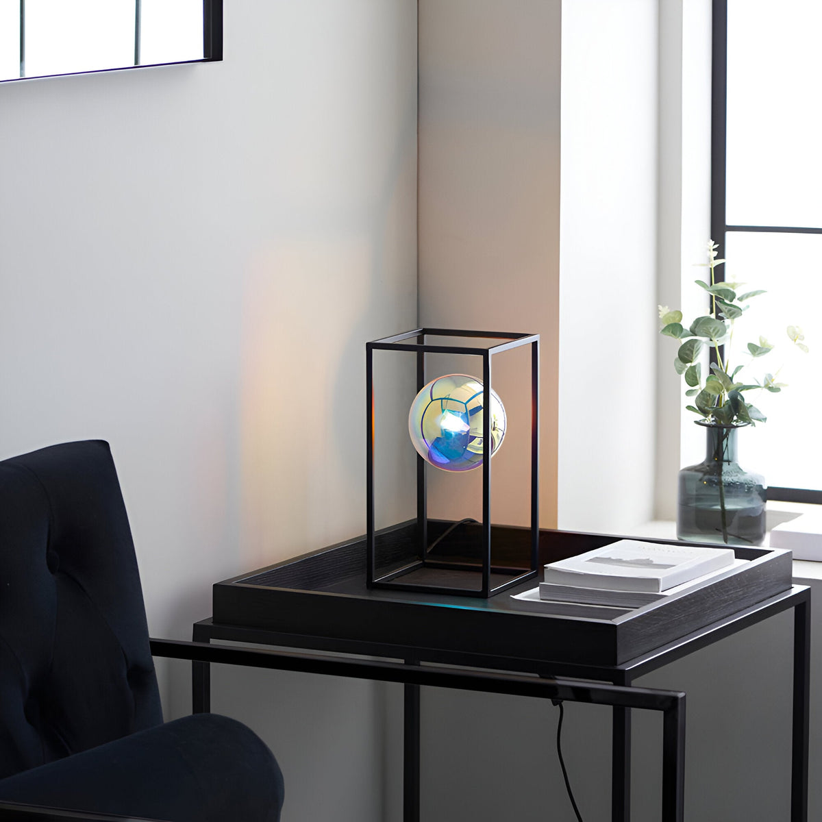 Brockwell Table Lamp in black with an open geometric frame and iridescent orb glass shade, shown on a side table in a living room setting.