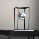 Brockwell Table Lamp in matt black with an open rectangular frame and iridescent orb glass shade lit within, shown on a dark table against a neutral background.