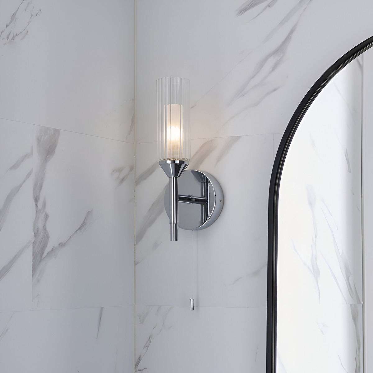 Endon Castbourne bathroom wall light in polished chrome with a ribbed clear glass torch shade, mounted on a marble wall beside an arched black-framed mirror.
