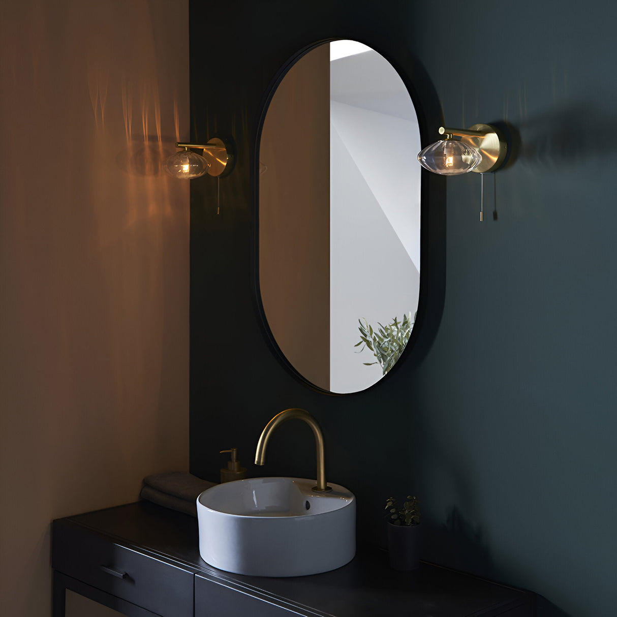 Carlmont Bathroom Wall Light in satin brass with ribbed glass shade and pull switch, shown either side of an oval mirror above a vanity sink.