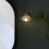 Carlmont Bathroom Wall Light in satin brass with ribbed glass shade and pull switch, mounted on a dark wall beside a curved mirror and greenery.