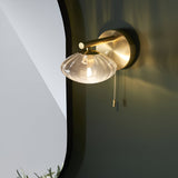 Carlmont Bathroom Wall Light in satin brass, with a rod arm holding a ribbed glass shade and pull cord switch, shown beside a mirror.