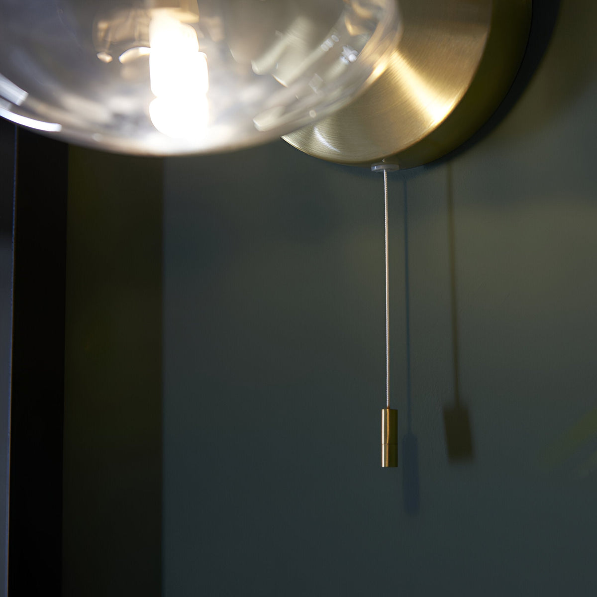 Carlmont Bathroom Wall Light in satin brass with round backplate, ribbed glass globe shade and brass pull cord switch against a plain wall.