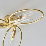 Close-up of Endon Hartley 3 light semi-flush ceiling light in brass, with oval rings holding clear glass cylinder shades and E14 lamps against a white ceiling.