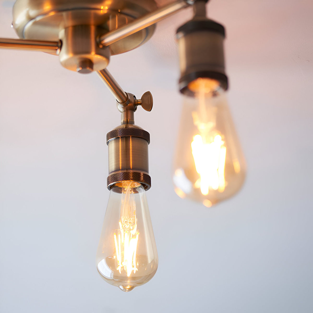 Close-up of two Endon Brackley semi-flush ceiling light arms in brass, each holding a lit E27 vintage-style teardrop filament bulb against a pale ceiling.