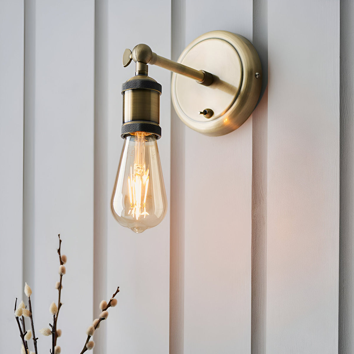 Brackley wall light in antique brass with an adjustable arm and exposed E27 bulb, mounted on a white panelled wall beside pussy willow stems.