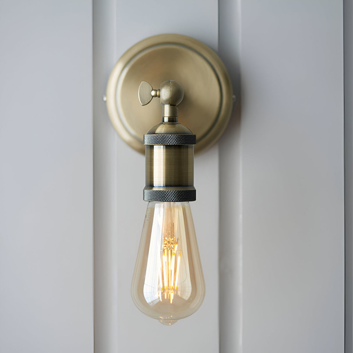 Antique brass Brackley wall light, industrial-style single E27 fixture with round backplate, adjustable knurled head, mounted on a white panelled wall.