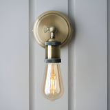 Antique brass Brackley wall light, industrial-style single E27 fixture with round backplate, adjustable knurled head, mounted on a white panelled wall.
