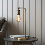 Brackley Table Lamp in brass with adjustable knuckle joint and exposed E27 bulb, shown on a side table beside a book in a living room setting.