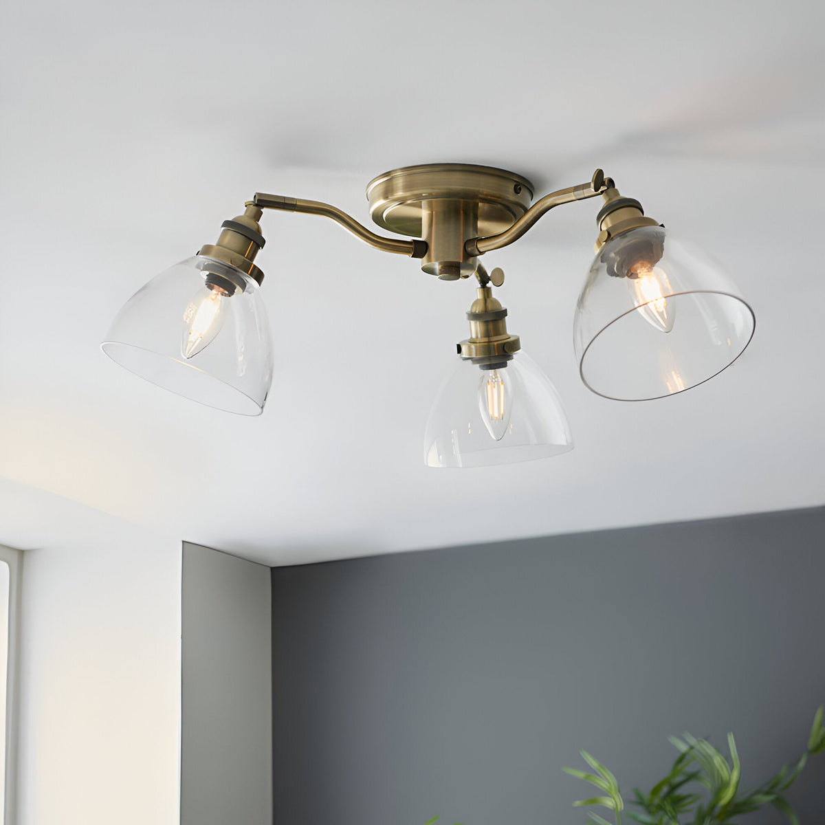 Wentworth 3 Light Semi-Flush Ceiling Light in antique brass with three angled arms and clear glass shades, mounted on a white ceiling above a grey wall.