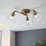 Wentworth 3 Light Semi-Flush Ceiling Light in antique brass with three angled arms and clear glass shades, mounted on a white ceiling above a grey wall.