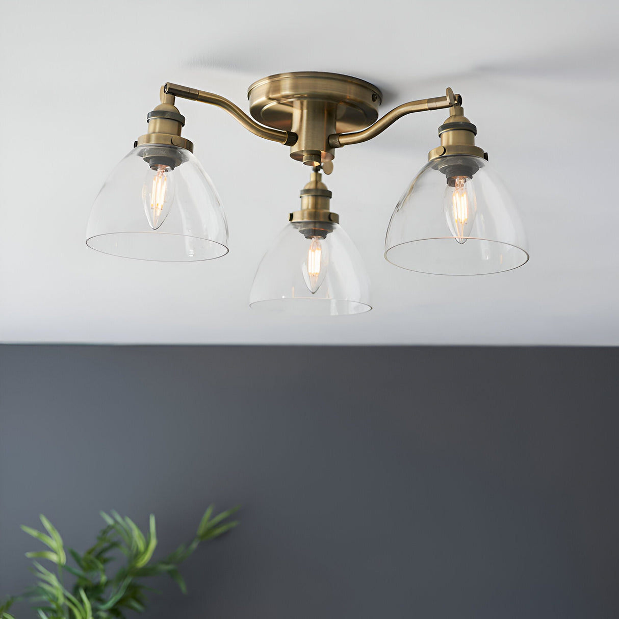Wentworth 3 Light Semi-Flush Ceiling Light in antique brass with three angled arms and clear dome glass shades, shown against a white ceiling and grey wall.