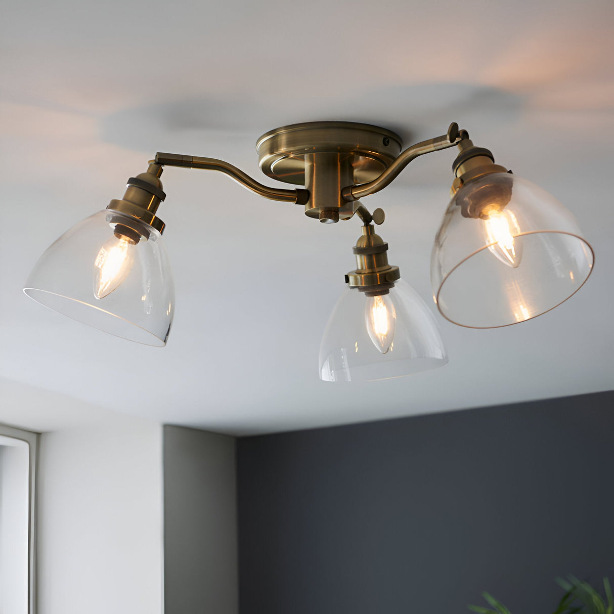 Wentworth 3 Light Semi-Flush Ceiling Light in antique brass with clear glass shades and visible bulbs, set against a white ceiling and dark grey wall.