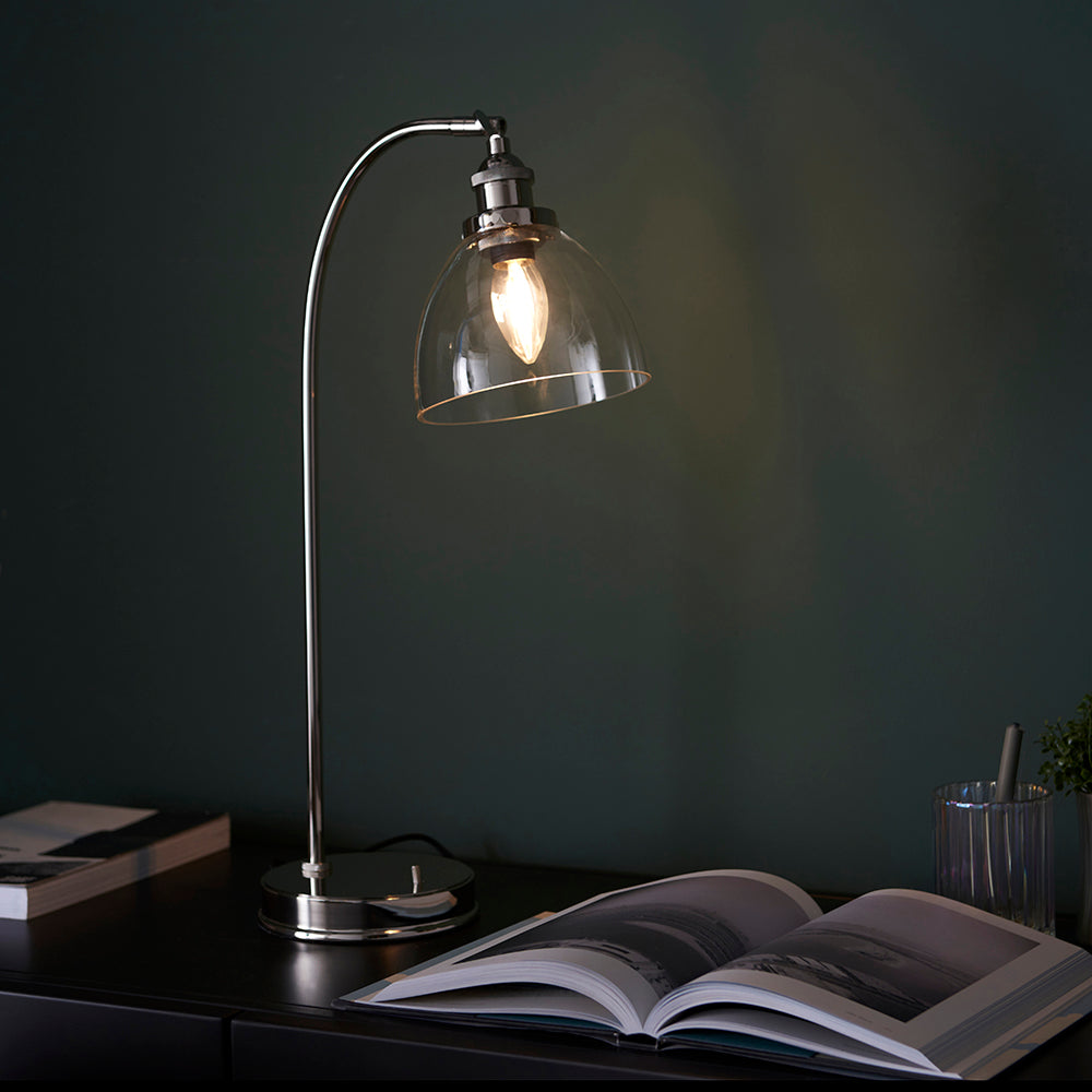 Endon Wentworth table lamp in nickel with an arched stem, clear glass dome shade and lit filament bulb, on a dark console beside an open book.