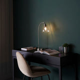 Endon Wentworth nickel table lamp with arched arm and clear glass shade lit on a black desk with books, beside a velvet chair in a dark room.