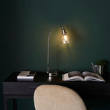 Endon Wentworth nickel table lamp lit on a black desk with an open book and pen pot, set against a dark green wall with a suede chair in front.