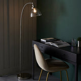Endon Wentworth nickel floor lamp with arched stem and clear glass shade beside a black desk and velvet chair in a dark green room.