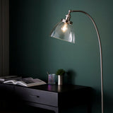 Endon Wentworth nickel floor lamp with arched arm and clear glass shade over a lit E27 bulb, beside a dark console table against a deep green wall.