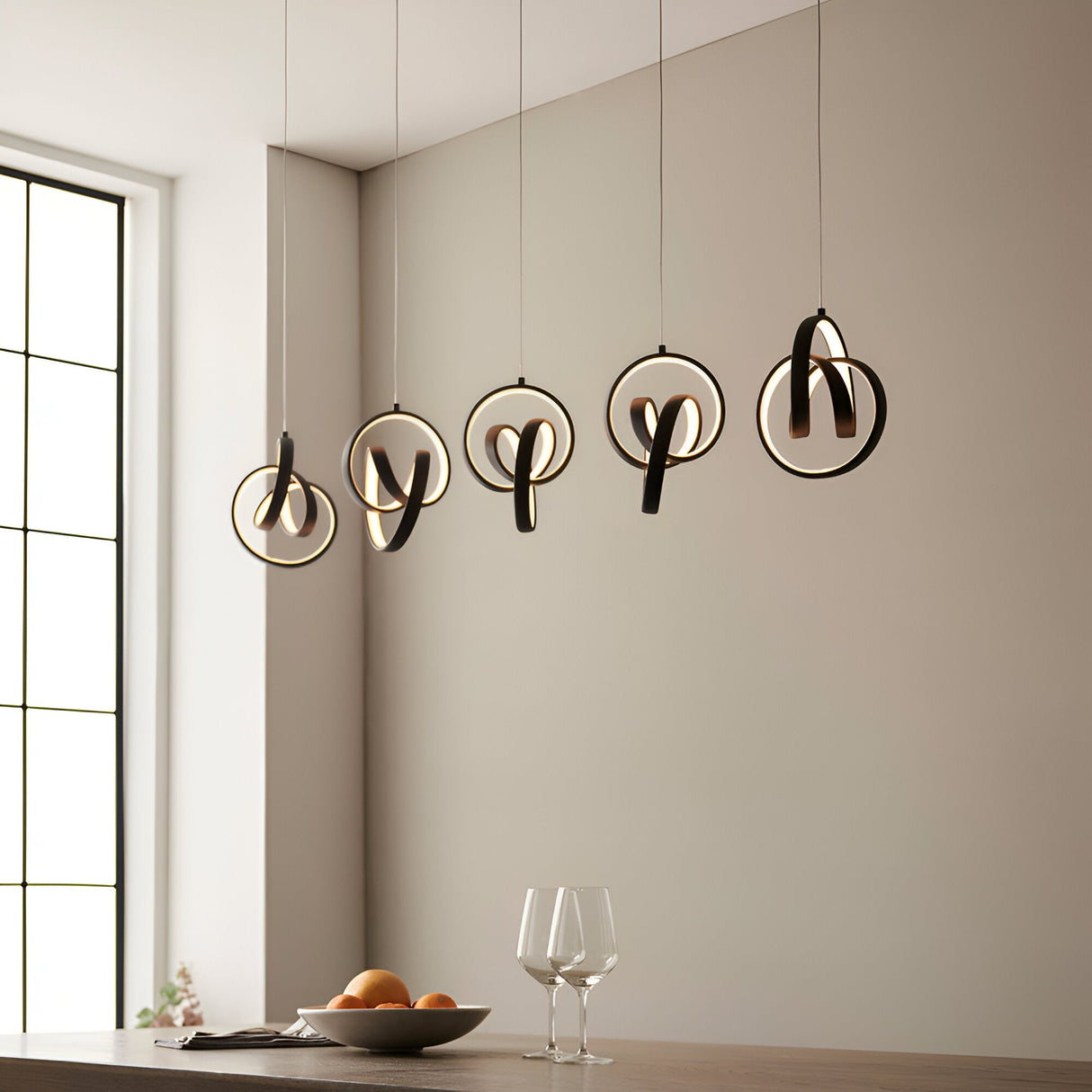 Bevwell 5 light linear pendant in matt black metal, looped LED rings suspended at varying drops above a dining table in a neutral room.