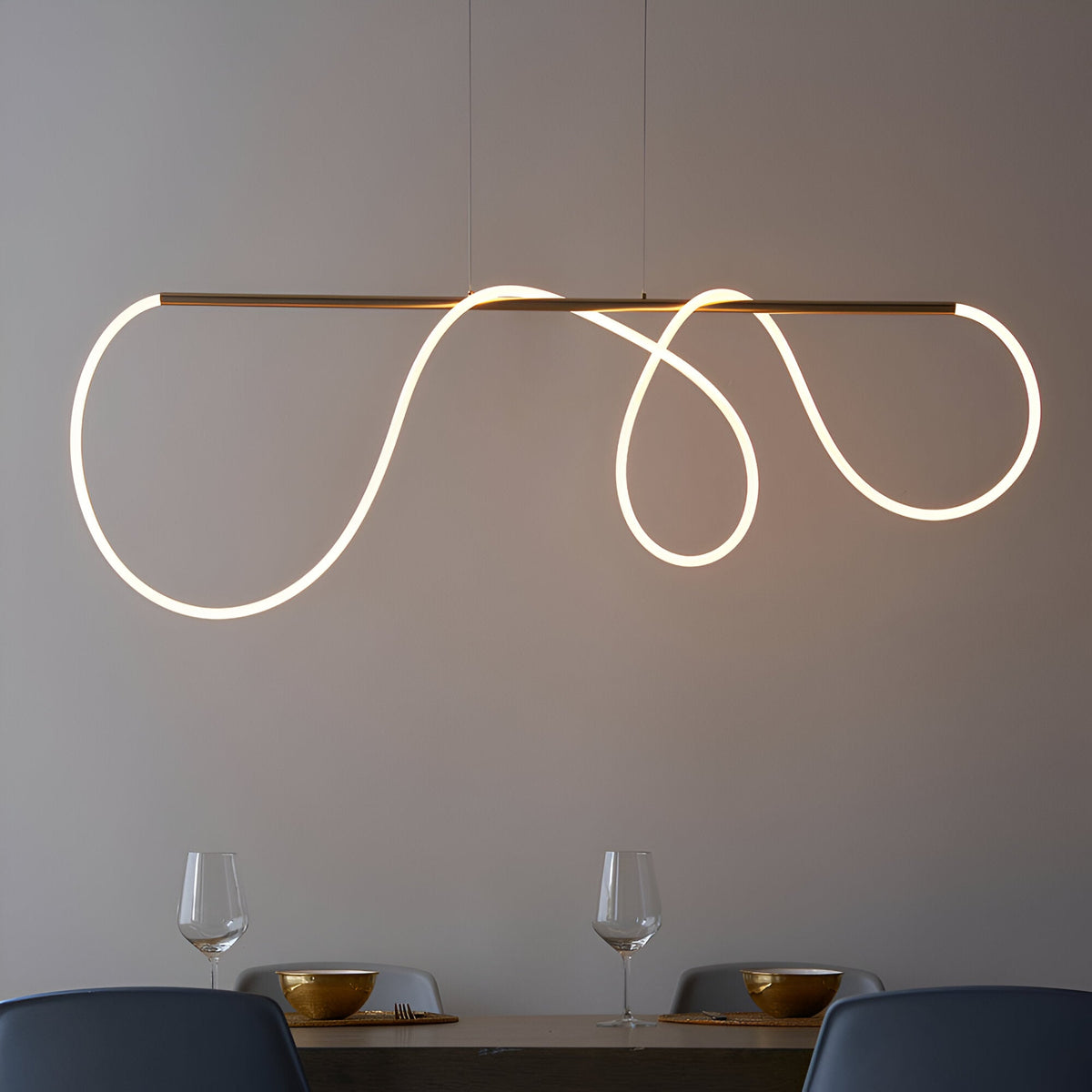 Aldley Linear Pendant in satin gold with a flexible LED tube curved into loops, suspended over a dining table with glassware and chairs below.