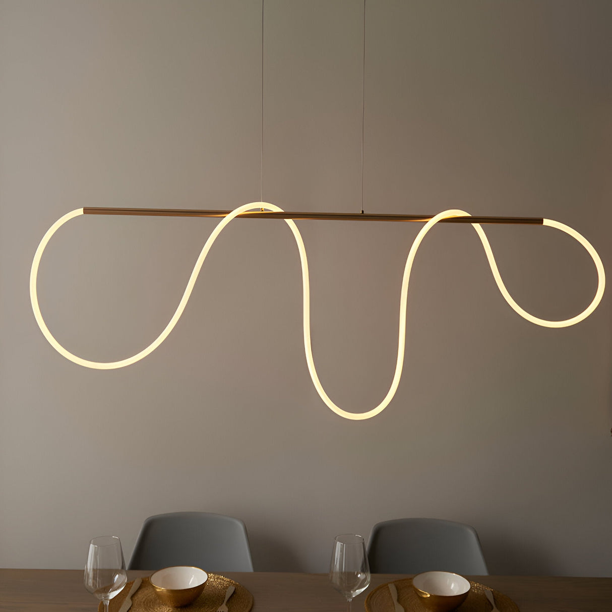Aldley Linear Pendant in satin gold, a horizontal bar with a flexible LED tube curving into wave shapes, hung above a dining table with grey chairs.