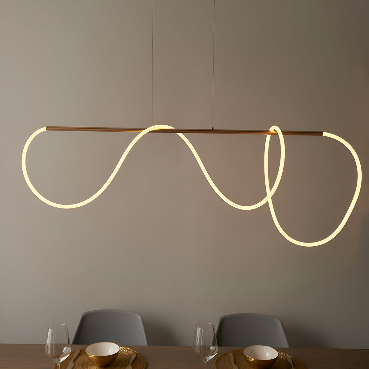 Aldley Linear Pendant in satin gold, a straight bar with a flexible warm-lit tube looping into curved waves, hung above a dining table with grey chairs.
