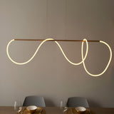Aldley Linear Pendant in satin gold, a straight bar with a flexible warm-lit tube looping into curved waves, hung above a dining table with grey chairs.