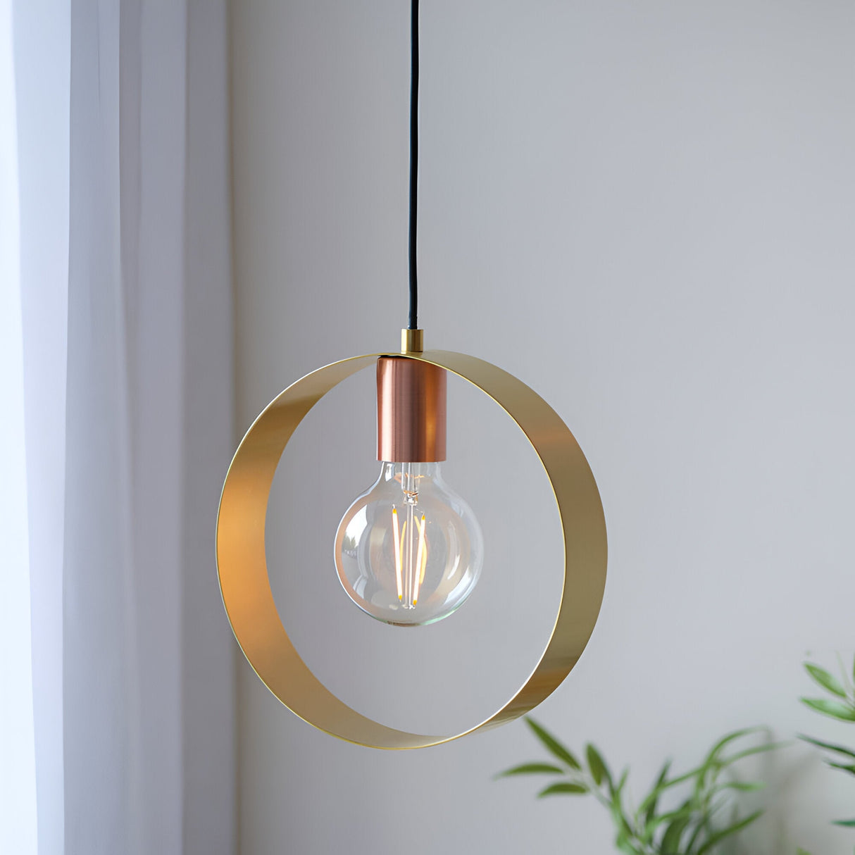 Brenmere Pendant Light with an open brass hoop frame, copper lamp holder and exposed globe bulb, hung against a light wall near curtains and greenery.