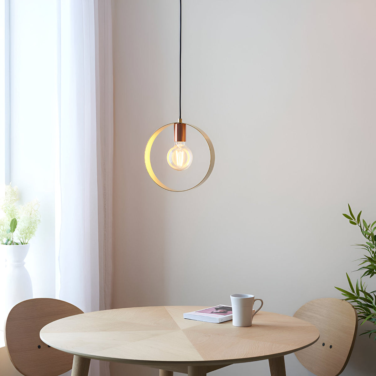Brenmere Pendant Light with brushed brass hoop frame and exposed filament bulb, hung above a round wood dining table in a light-toned room.