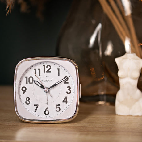 Square white alarm clock with silver trim, bold black numerals and sweep hands, shown on a wooden surface beside decorative home accessories.