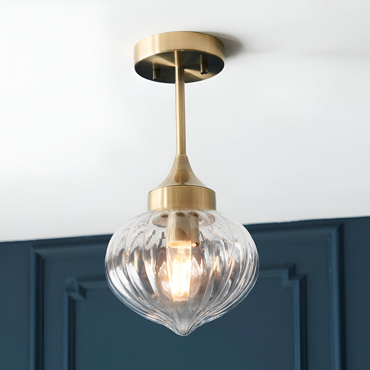 Endon Aldridge semi-flush ceiling light in antique brass with a ribbed clear glass teardrop shade, mounted below a white ceiling by a dark blue panelled wall.