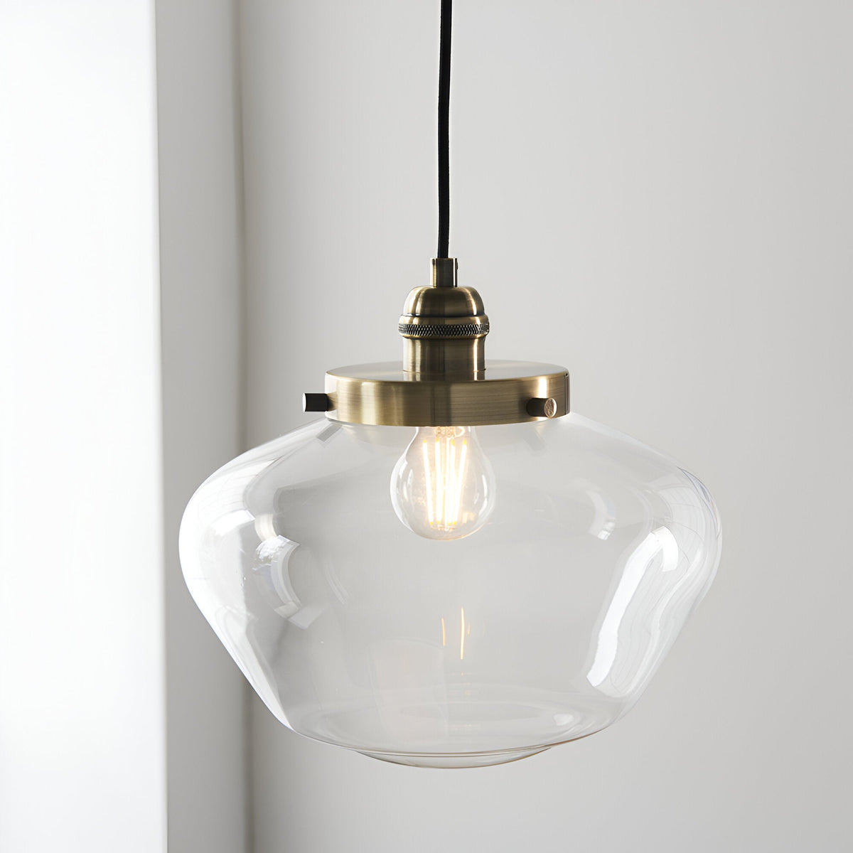 Kedleston Pendant in brass with a clear rounded glass shade and exposed E27 bulb, hung against a plain light grey wall.