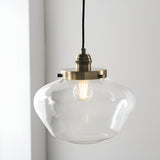 Kedleston Pendant in brass with a clear rounded glass shade and exposed E27 bulb, hung against a plain light grey wall.