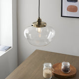 Kedleston Pendant in brass with a clear glass schoolhouse shade, hung above a wood dining table beside a framed print on a light wall.