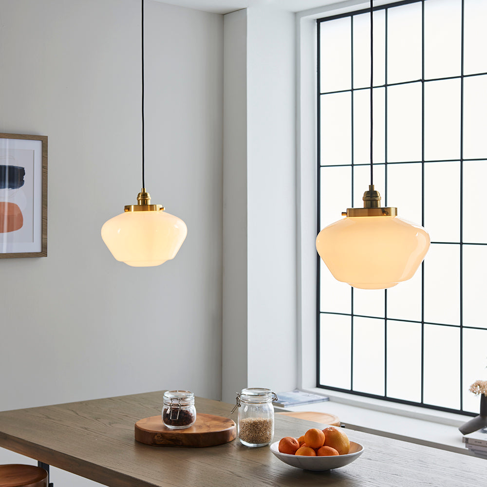 Endon Kedleston pendant lights with brass fittings and opal glass schoolhouse shades on black flex, hung over a wooden dining table by a windowed kitchen wall.